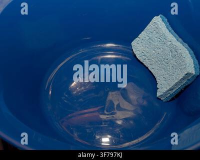 Close up of a light blue sponge resting inside a dark blue plastic bucket or basin with soapy water Stock Photo