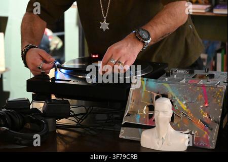 A horizontal photograph capturing a close-up of a person's hands operating a DJ turntable, placing a vinyl record, with a watch on the wrist, a pendant around the neck, and a small white bust on the table. Stock Photo