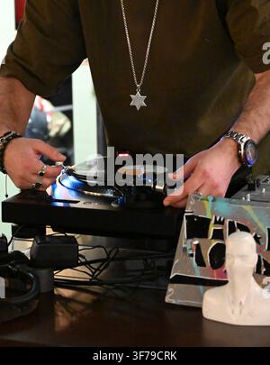 A horizontal photograph capturing a close-up of a person's hands operating a DJ turntable, placing a vinyl record, with a watch on the wrist, a pendant around the neck, and a small white bust on the table. Stock Photo