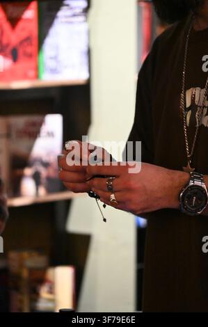A close-up photograph capturing a DJ's hands, adorned with silver rings and a watch, meticulously adjusting the delicate stylus (needle) on a vinyl record player cartridge. The turntable is playing a record, and blurred vinyl shelves are in the background. Stock Photo