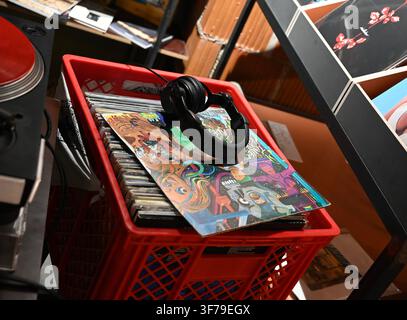 A high-angle photograph capturing a pair of black headphones resting on top of a colorful vinyl record sleeve inside a bright red plastic crate filled with many other records, in a record store setting. Stock Photo