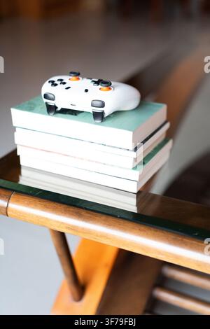 White gaming controller resting on a stack of books atop a wooden coffee table with glass top in a Stock Photo