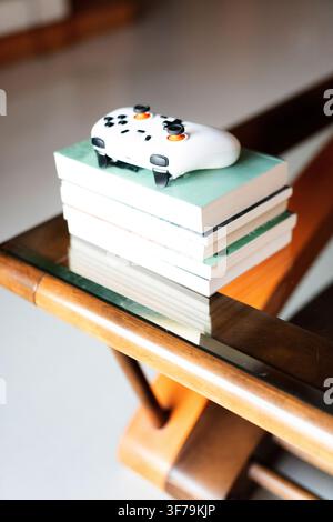 White gaming controller resting on a stack of books atop a mid-century modern glass-top wooden Stock Photo