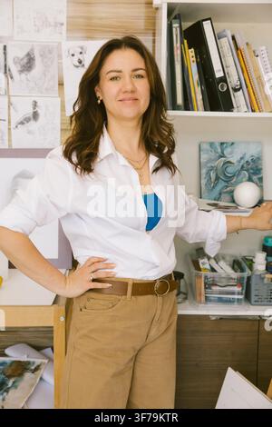 Vertical portrait of a smiling female artist standing in her creative studio surrounded by paintings, sketches and art supplies. Authentic lifestyle i Stock Photo