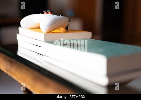 White PlayStation controller resting on a stack of books atop a wooden table in a cozy indoor Stock Photo