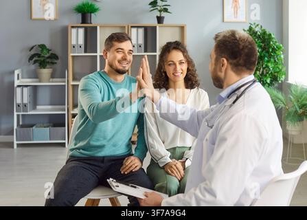 Male family doctor giving high five with a young happy married couple sitting in medical clinic. Stock Photo