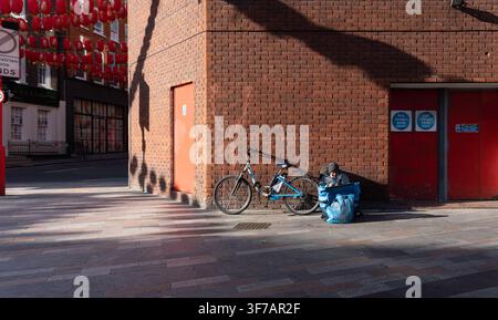 A Stuart delivery rider checks their phone while waiting for orders by a red brick wall on Macclesfield Street in London’s Chinatown. Stock Photo