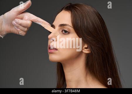 Surgeon adjusting adhesive tape on the nose of a female patient after a rhinoplasty procedure. Doctor examining nasal taping on a young woman followin Stock Photo