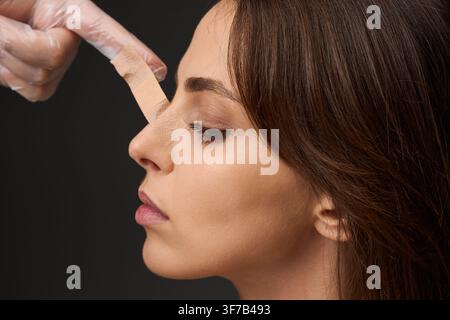 Surgeon adjusting adhesive tape on the nose of a female patient after a rhinoplasty procedure. Doctor examining nasal taping on a young woman followin Stock Photo
