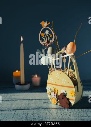 Decorative Easter arrangement featuring an egg shaped basket filled with colorful spring decorations surrounded by candles and festive branches on a b Stock Photo