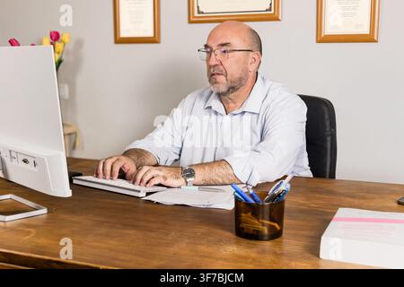 Mature businessman typing at a computer in a professional office with paperwork, pen holder and framed certificates. Great for business, productivity, Stock Photo