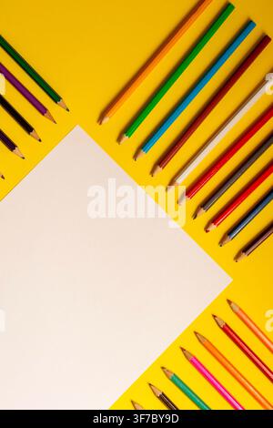 A blank sheet of paper sits on a yellow surface. Around it are many colored pencils arranged neatly. The setting shows preparation for school activiti Stock Photo