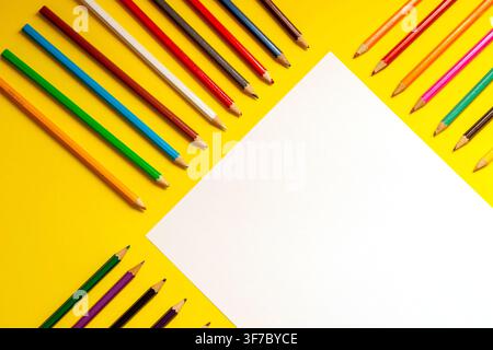 Colorful pencils surround a blank sheet of paper on a yellow table. This setup is ready for school activities. It shows a cheerful work area for stude Stock Photo