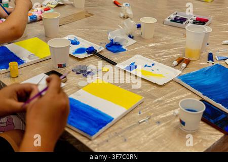 Students are engaged in an art activity in their classroom as they paint on paper and use various colors of paint. The setting is lively with supplies Stock Photo