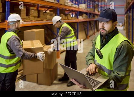 Warehouse manager using laptop while team handles boxes during stock inventory Stock Photo