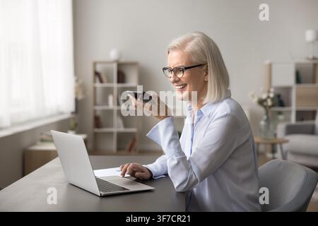 Smart older lady sitting by laptop dictate voicemail on cell Stock Photo