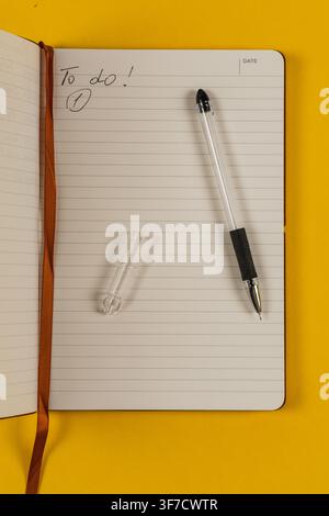 A notebook lies open on a yellow desk with a pen next to it. The page shows a to-do list. Stock Photo