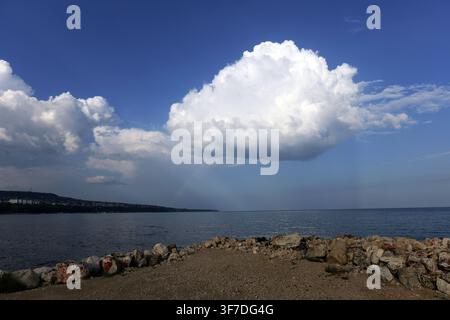 Large cumulus cloud dominating a deep blue sky above the calm sea, with a distant coastline and a rocky barrier in the foreground Stock Photo