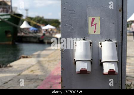 Industrial electrical sockets on a metal pole featuring a lightning bolt symbol. Offering shore power for boats and ships docked at the bustling harbo Stock Photo