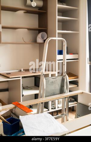 Setting up wooden cabinetry at a home improvement site using a step ladder for reach. Workspace features multiple shelves, tools, and building materials visible. New kitchen concept.  Stock Photo