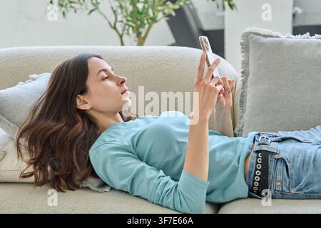 Serious young woman using smartphone lying on sofa at home Stock Photo