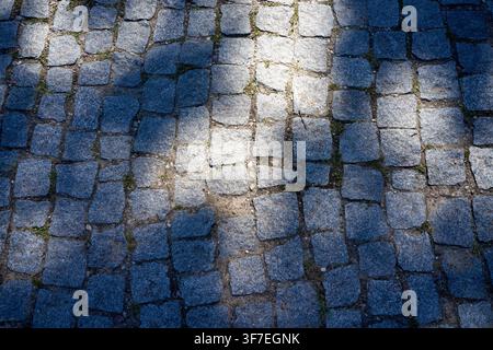Cobbled street bathed in dappled sunlight. Shadows stretch across weathered stone tiles. Green moss creeps between each stone. Patterned light adds te Stock Photo