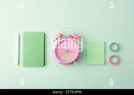 Minimal pastel workspace featuring pink alarm clock, green notebook, pen, sticky note and decorative tape on mint background. Back to school concept. Stock Photo