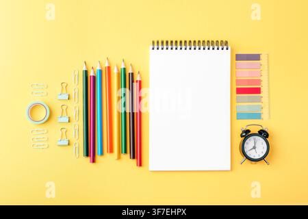 Stationery composition with colored pencils, blank notebook, ruler, paper clips and alarm clock on bright yellow surface. Back to school concept. Top Stock Photo