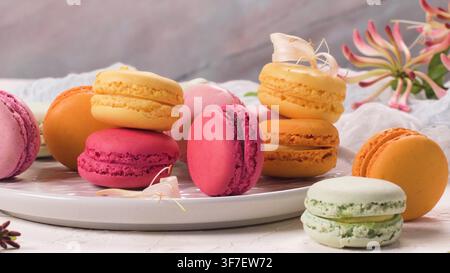 An assortment of vibrant French macarons rests on a ceramic plate, accompanied by delicate honeysuckle blossoms. Soft natural lighting highlights the Stock Photo
