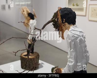 Two contemporary wooden sculptures displayed in an art gallery setting. The foreground sculpture depicts a stylized human figure wearing a white Stock Photo