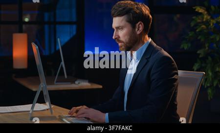 Serious concentrated adult Caucasian business man working with computer at late night evening in office workplace businessman male executive boss work Stock Photo