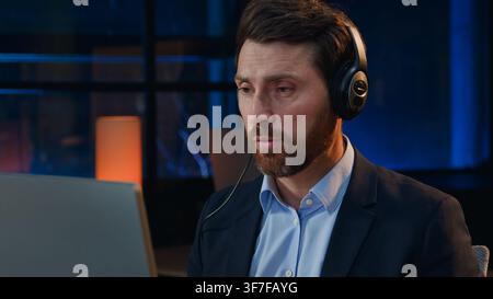 Concentrated Caucasian middle-aged business man in headphones talking video call at night evening office mature businessman male in headset consultant Stock Photo