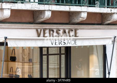 ITALY, VENICE, 11 JULY 2025:A Versace store facade with white awning, large signage, and handbags displayed in the window is shown beneath an ornate b Stock Photo
