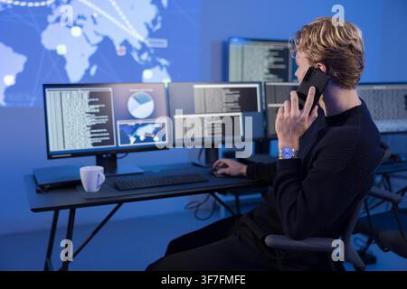 A cybersecurity analyst monitors multiple computer screens displaying network data, logs, and maps. The environment appears to be a SOC, and the indiv Stock Photo