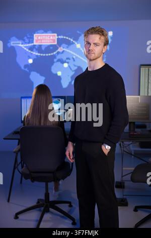 Two cybersecurity professionals work actively in a secure operations center equipped with advanced displays. A young man stands confidently while a fe Stock Photo
