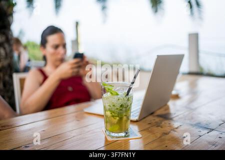 focused young digital nomad woman doing remote work on laptop at outdoor terrace cafe with a mojito drink, nomad lifestyle concept Stock Photo
