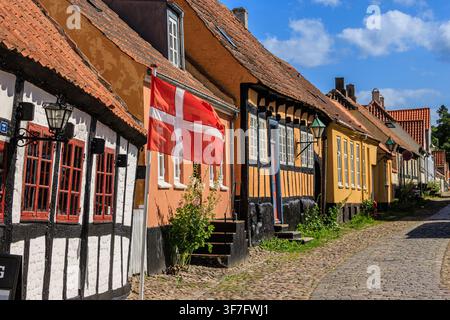 Sunlit historic Ebeltoft street with colourful half-timbered houses, a waving Denmark flag and cobblestone pavement in warm summer light Stock Photo