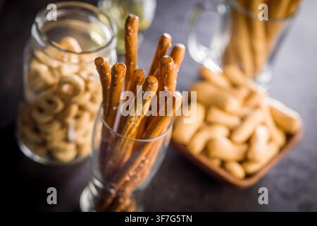 Close-up of salted pretzel sticks served in a glass with jars and a bowl of assorted snacks on a dark tabletop, evoking casual gatherings, party appet Stock Photo