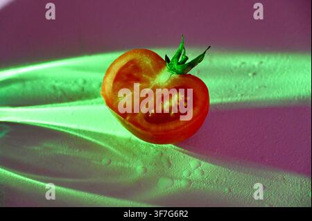 Close-up commercial shot of a fresh ripe red tomato half with a green stem, positioned centrally under neon green light rays against a dark purple and pink textured background. Suitable for conceptual food photography, modern culinary design, and creative advertising. Stock Photo