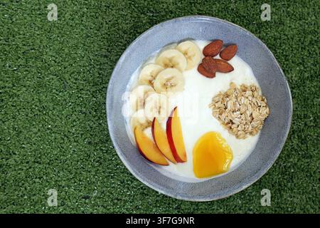 Yogurt bowl filled with banana slices, peach wedges, almonds, granola, and honey on a green background, representing a nutritious and refreshing meal Stock Photo