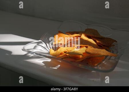 Savory corn tortilla chips are presented in a decorative glass bowl, catching a strong diagonal beam of sunlight that creates distinct shadows on a wh Stock Photo