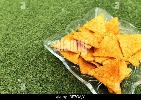Nacho chips with seasoning filling a clear glass bowl, creating a savory snack display against a vibrant green artificial grass background, perfect fo Stock Photo