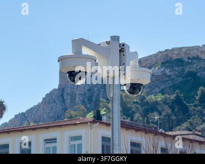 Two dome security cameras on a metal pole monitor buildings below a rocky hillside in Monastiraki, Athens, Greece, blue sky Stock Photo