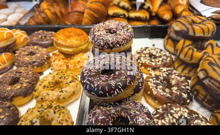 Stacks of ring doughnuts coated in chocolate, caramel, nuts and rainbow sprinkles create an abundant sweet pastry selection on trays Stock Photo
