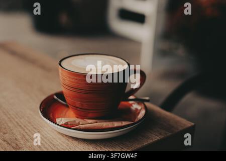 A creamy coffee served in a red cup and saucer sits on a wooden cafe table, photographed in soft natural light with warm tones, shallow depth of field Stock Photo