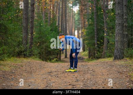 Man in athletic apparel taking a break from exercising on a forest path, checking his progress and location on a smartphone during an outdoor workout, focusing on health and personal bests Stock Photo