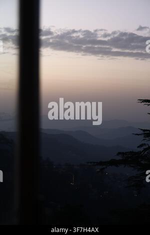 Silhouette of pine tree branch overlooking soft pastel sunset sky and layered blue mountain ridges in distance. Stock Photo