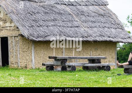 Thatched-roof mud cottages sit on a lush green field beside wooden picnic tables, evoking rural history, simplicity and outdoor leisure in a reconstru Stock Photo