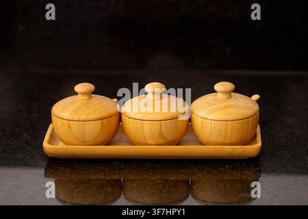 Three round bamboo jars with lids on a matching rectangular tray. Set on a dark, reflective kitchen surface. Minimalist design showcasing natural wood Stock Photo