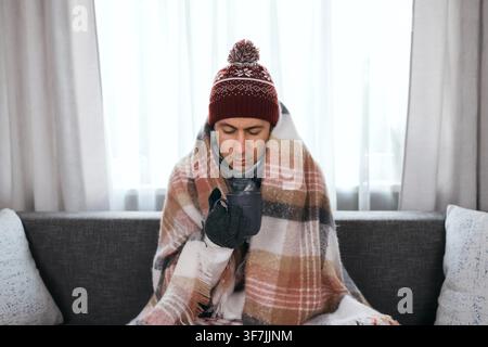 Freezing man wrapped in a blanket holds a warm mug at home. High heating costs and the energy crisis cause indoor cold weather, highlighting a severe Stock Photo